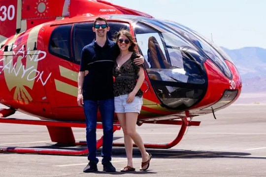 Couple smiling in front of a red helicopter on a sunny day.
