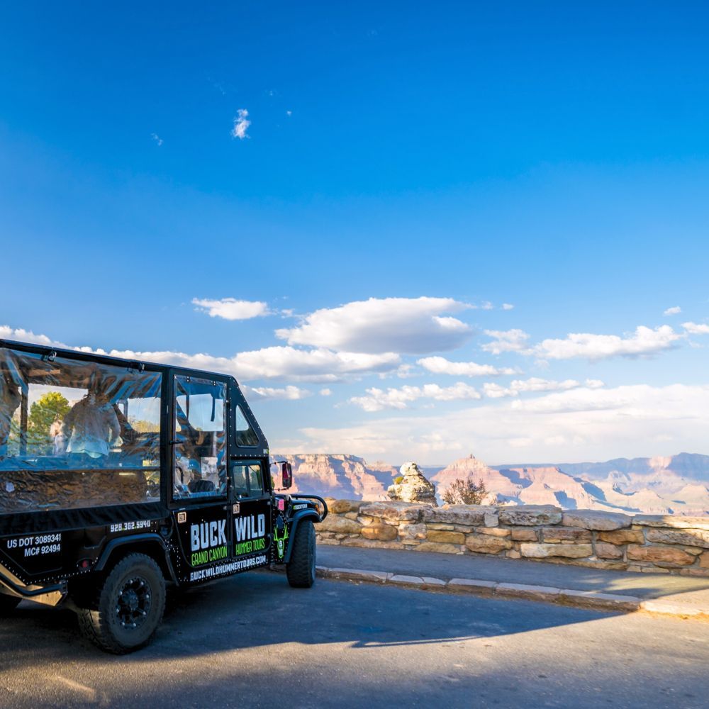 Tour jeep parked at a scenic overlook with a view of the Grand Canyon under a clear blue sky.